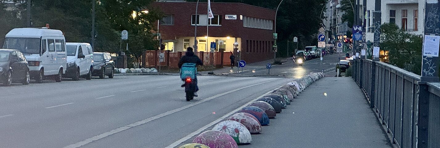 Auf dem Bild ist ein Lieferdienst-Fahrer mit einer blauen Wolt-Transport-Box auf dem Fahrrad zu sehen. Er fährt auf einer großen Straße in dämmrigem Licht. 