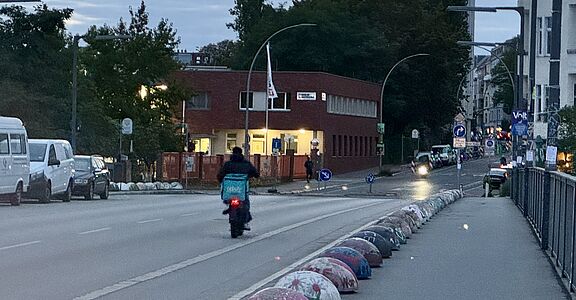 Auf dem Bild ist ein Lieferdienst-Fahrer mit einer blauen Wolt-Transport-Box auf dem Fahrrad zu sehen. Er fährt auf einer großen Straße in dämmrigem Licht. 