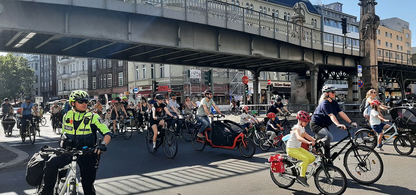Kidical Mass Schöneberg 21. September 2024 Eine Gruppe Erwachsener und Kinder fahren auf ihren Fahrrädern unter einer Brücke auf der Fahrbahn der Bülowstraße in Schönberg. Im Vordergrund ist eine Fahrradpolizistin zu sehen.