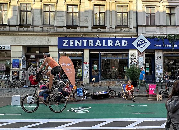 PARKing Day 2024 ADFC Berlin Ein Zweirad fährt über einen grünen Pop-Up Radweg, Menschen auf Liegestühlen im Hintergrund