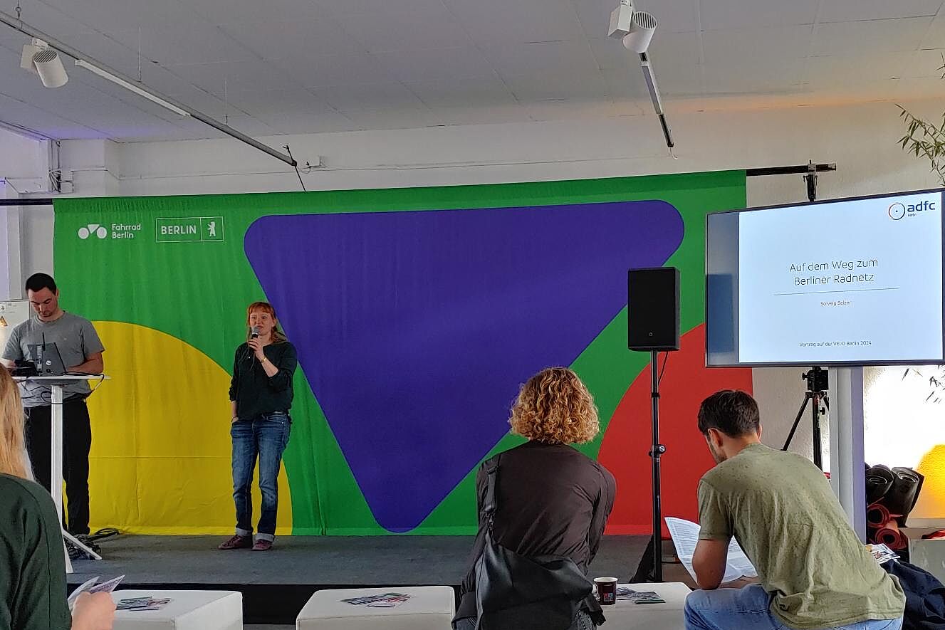 VELO 2024 Menschen sitzen auf weißen Hockern und hören einer Referentin auf dem Podium zu