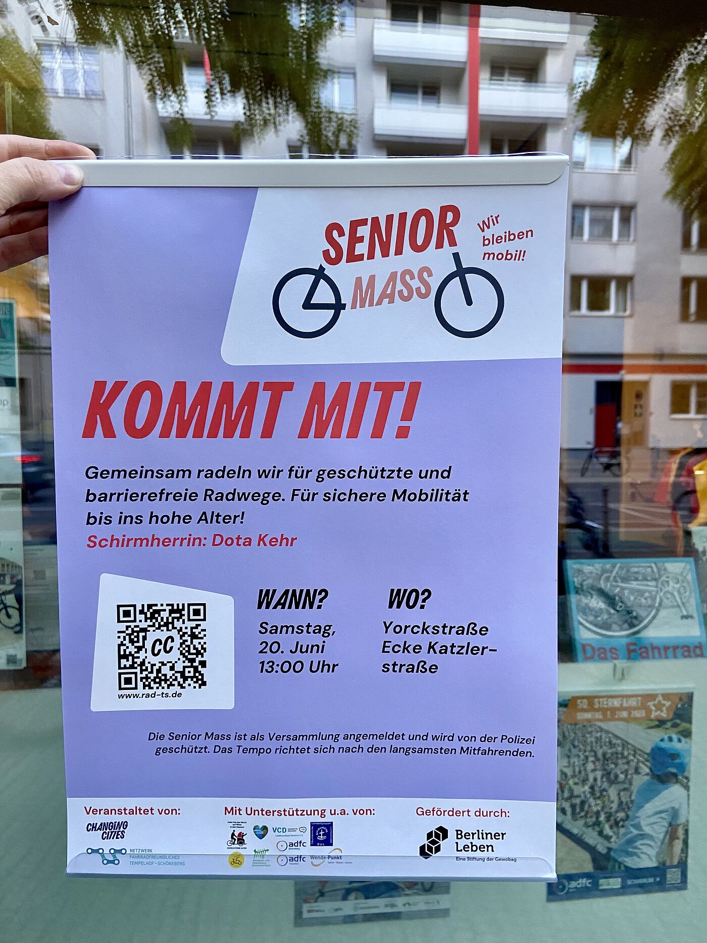 Die Senior Mass am 20.06.2026 wird mit einem Plakat im Schaufenster des ADFC Berlin VeloKiez in der Möckernstraße beworben. Plakat von Changing Cities wird vor einer Schaufensterscheibt des ADFC Berlin VeloKiez hochgehalten