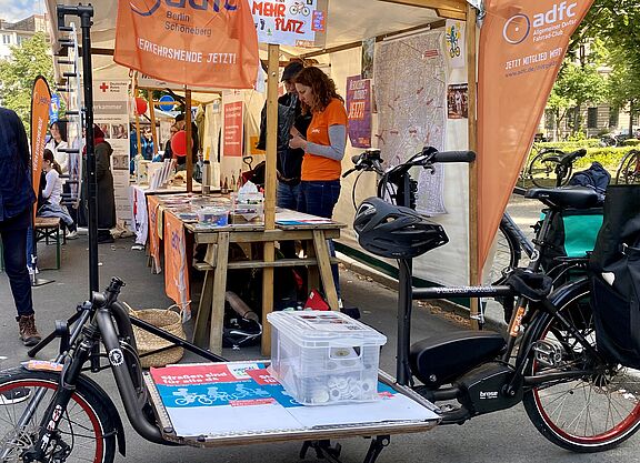 Stand des ADFC Schöneberg und Tempelhof beim Nachbarschaftsfest am Schöneberger Rathaus am 24.05.2025 mit Lastenrad e-Harry Stand des ADFC Schöneberg und Tempelhof beim Nachbarschaftsfest am Schöneberger Rathaus am 24.05.2025. Im Vordergund ist ein e-Lastenrad zu sehen. Auf der Ladefläche befindet sich Material für den Stand. Es sind Fahnen, Plakate und Beachflags des ADFC Berlin in Orangetönen zu sehen, und Personen, die den Stand betreuen.