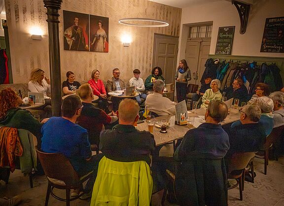 Podium_Wahlveranstaltung_ADFC_2026 Landtagskandidat*innen sitzen nebeneinander auf einer Bank im Kaffeehaus, davor aufmerksam zuhörende Menschen aus dem Publikumtgliedern und Interessierten im Kaffeehaus Gutmann