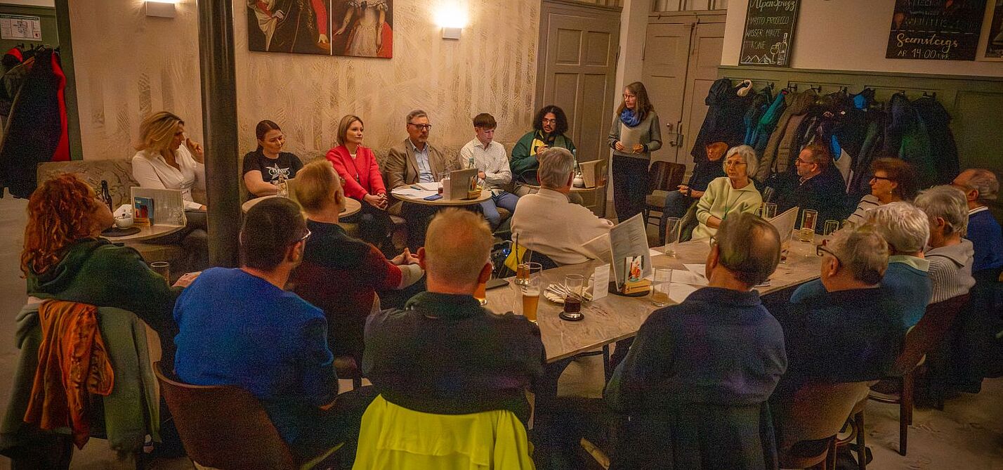 Podium_Wahlveranstaltung_ADFC_2026 Landtagskandidat*innen sitzen nebeneinander auf einer Bank im Kaffeehaus, davor aufmerksam zuhörende Menschen aus dem Publikumtgliedern und Interessierten im Kaffeehaus Gutmann