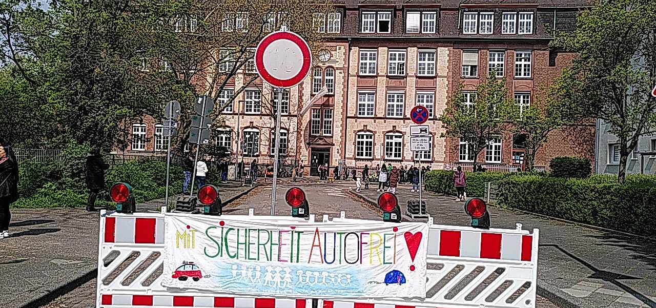 Schulstraße RH 2023 Absperrung Das Bild zeigt die Sperre an der Schulstraße mit einem von Kindern gemalten Transparent mit der Aufschrift 'Mit Sicherheit Autofrei'