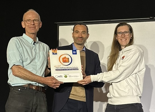 Nora Müller (links) vom ADFC SH übergibt die Plakette "Fahrradfreundliche Schule" an Mathias Kadow-Leder (Schulleiter Holstenschule) und Christian Wagner (Fahrradbeauftrager Holstenschule Neumünster)