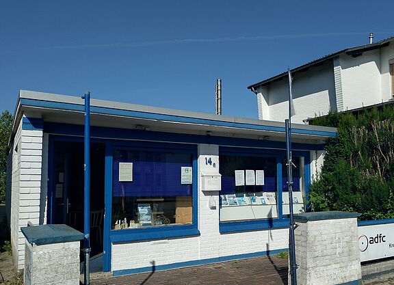 Infoladen Ein weißer, gemauerter Bungalow mit blauen Tür- und Fensterrahmen.