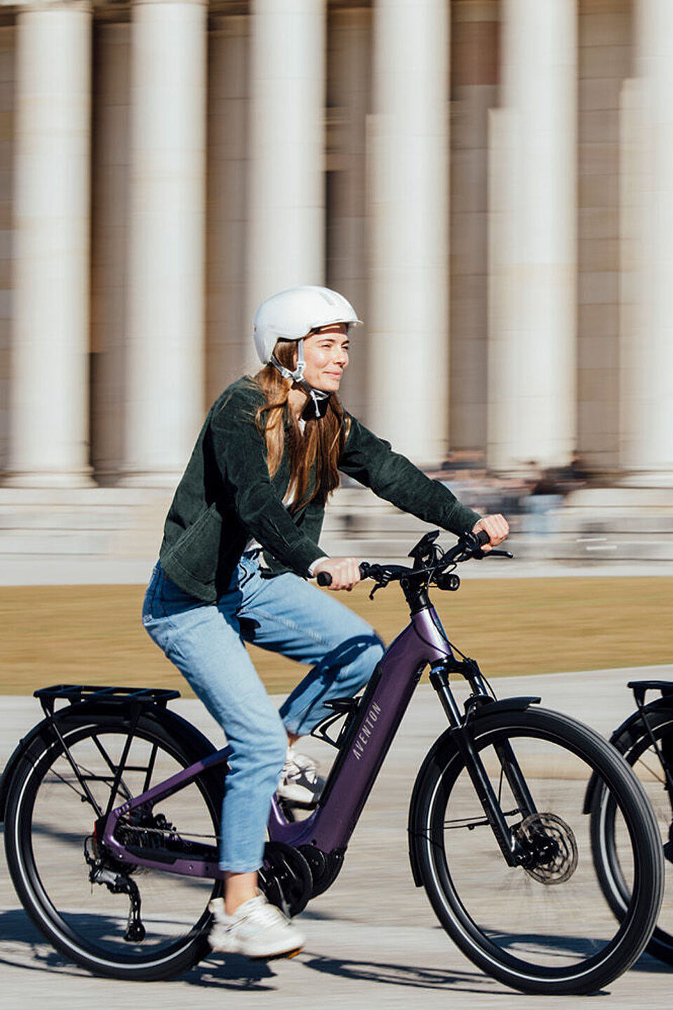 Aventon Zwei Menschen fahren mit Helmen auf Elektrofahrrädern an einem großen Gebäude mit Säulen vorbei.