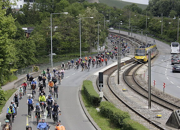 Große Gruppe von Radfahrenden auf einer breiten Straße neben Bahngleisen in Stuttgart. Die Teilnehmenden tragen bunte Sportkleidung und Fahrradhelme. Links verläuft parallel eine Stadtbahn auf Gleisen, rechts ist eine Autobahn mit wenigen Fahrzeugen zu sehen.