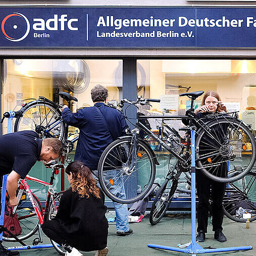 Mehrere Menschen reparieren Fahrräder auf Ständern vor einem Gebäude mit ADFC‑Berlin‑Schild.