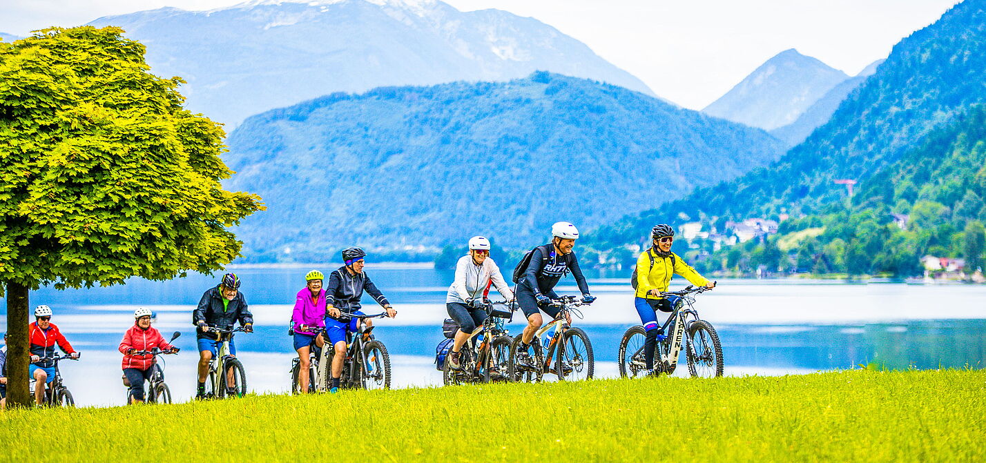 Radtour in der Gruppe am See Gruppe von Radfahrenden auf Wiese am See mit Bergpanorama im Hintergrund.