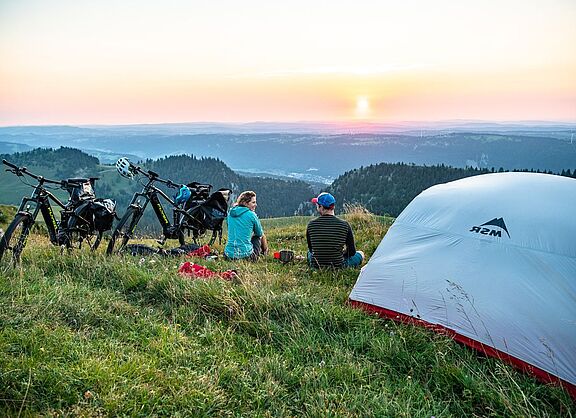 Zwei Radreisende sitzen in der Dämmerung neben einem Zelt im Grünen und blicken auf einen Höhenzug in der Ferne. 