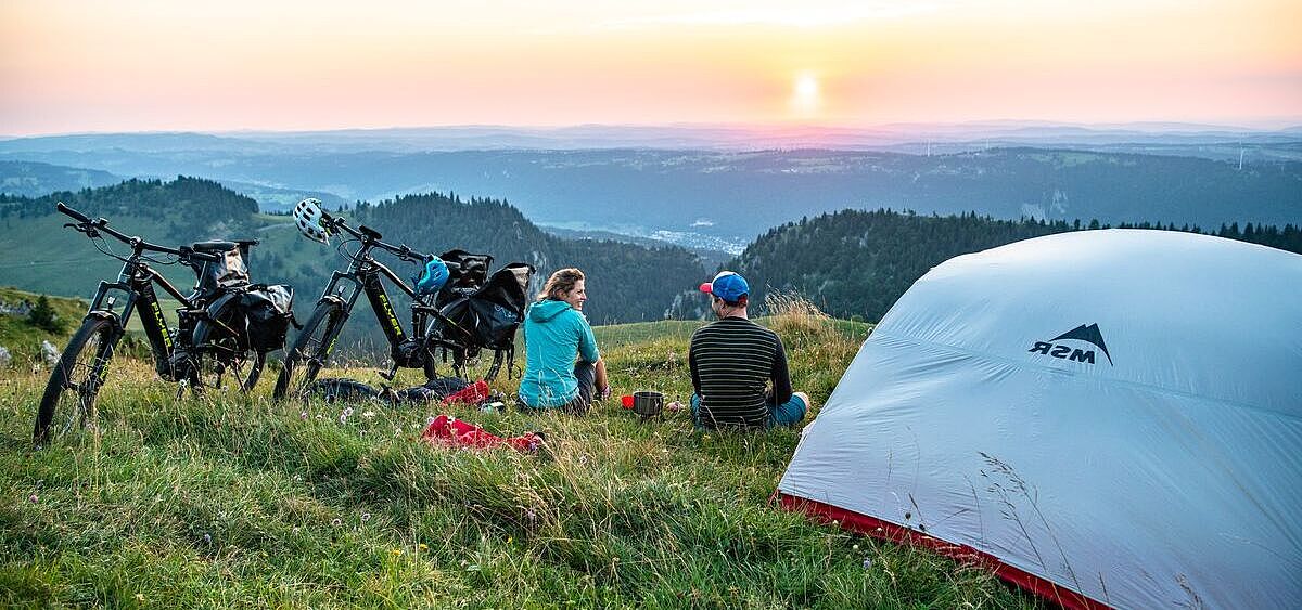 Zwei Radreisende sitzen in der Dämmerung neben einem Zelt im Grünen und blicken auf einen Höhenzug in der Ferne. 