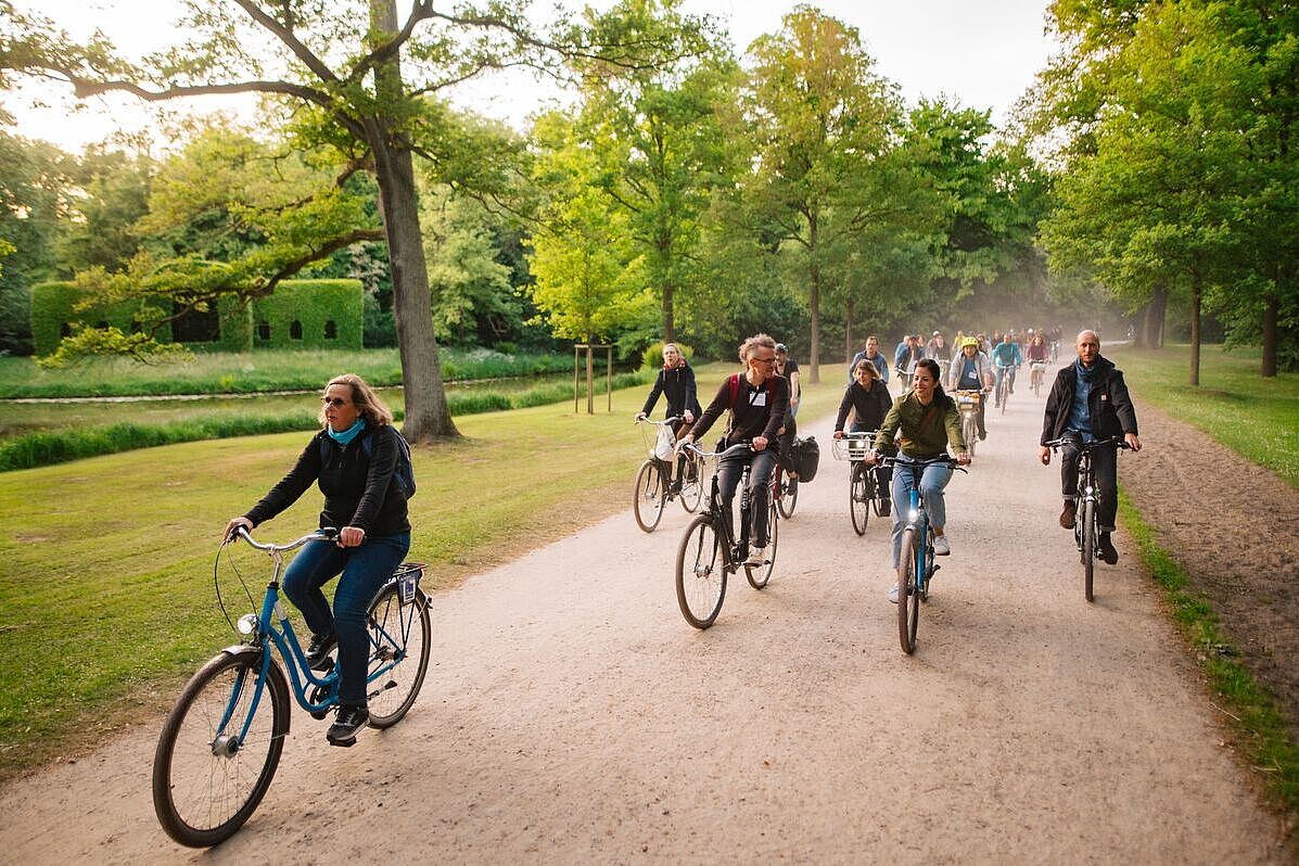 Eine Gruppe Radfahrender auf einem breiten Parkweg, umgeben von Bäumen und Rasenflächen. 