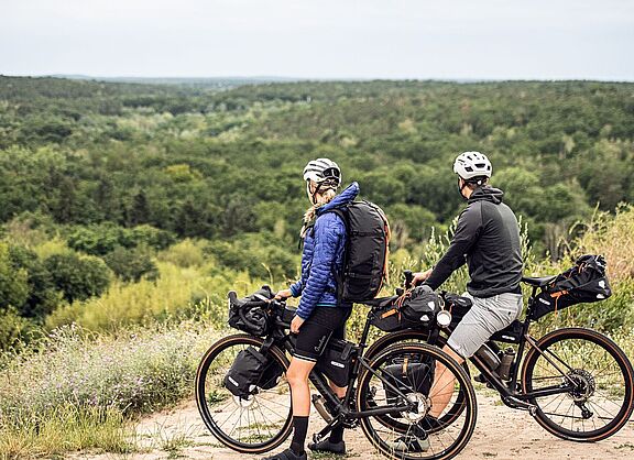 Zwei Radfahrende mit Gravelbikes und Gepäck stehen auf einer Anhöhe und blicken auf einen weiten Wald. 