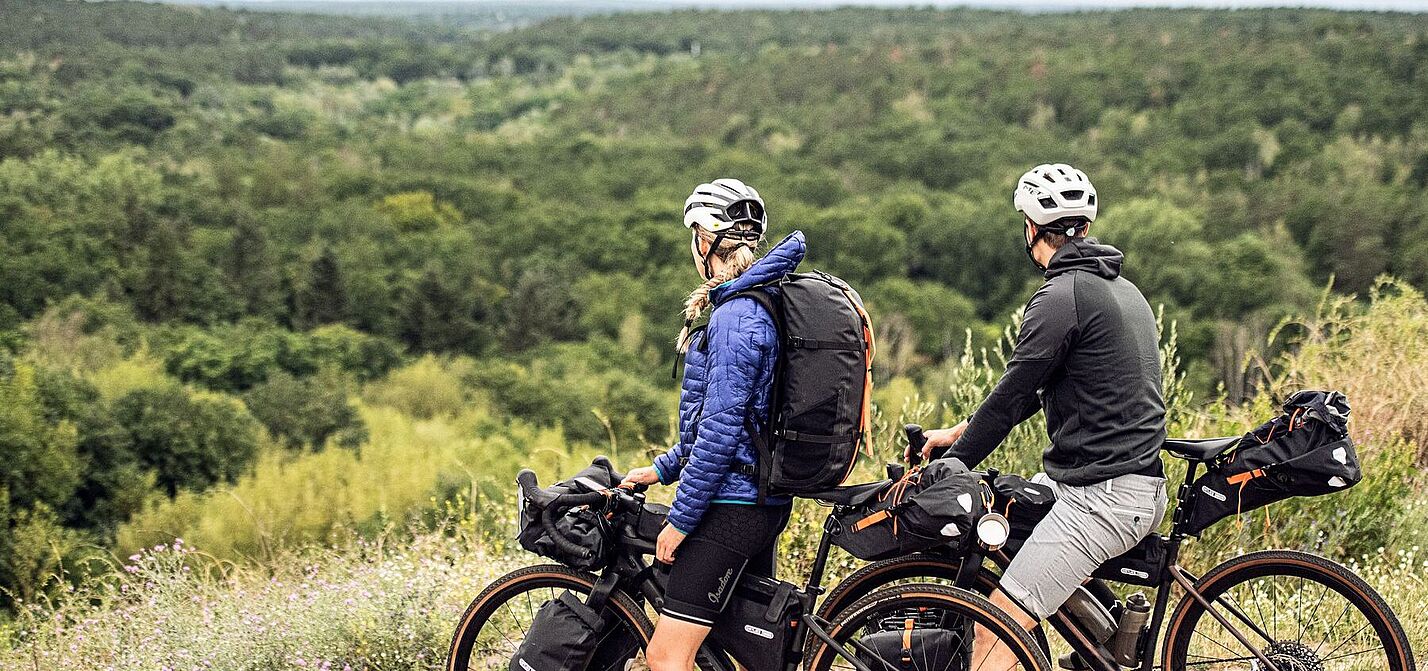 Zwei Radfahrende mit Gravelbikes und Gepäck stehen auf einer Anhöhe und blicken auf einen weiten Wald. 