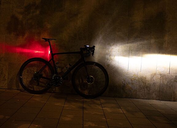 Ein Fahrrad steht in der Dunkelheit an einer Wand, mit am Lenker montiertem, eingeschalteten Scheinwerfer und an der Sattelstütze montierten, leuchtenden Rücklicht.