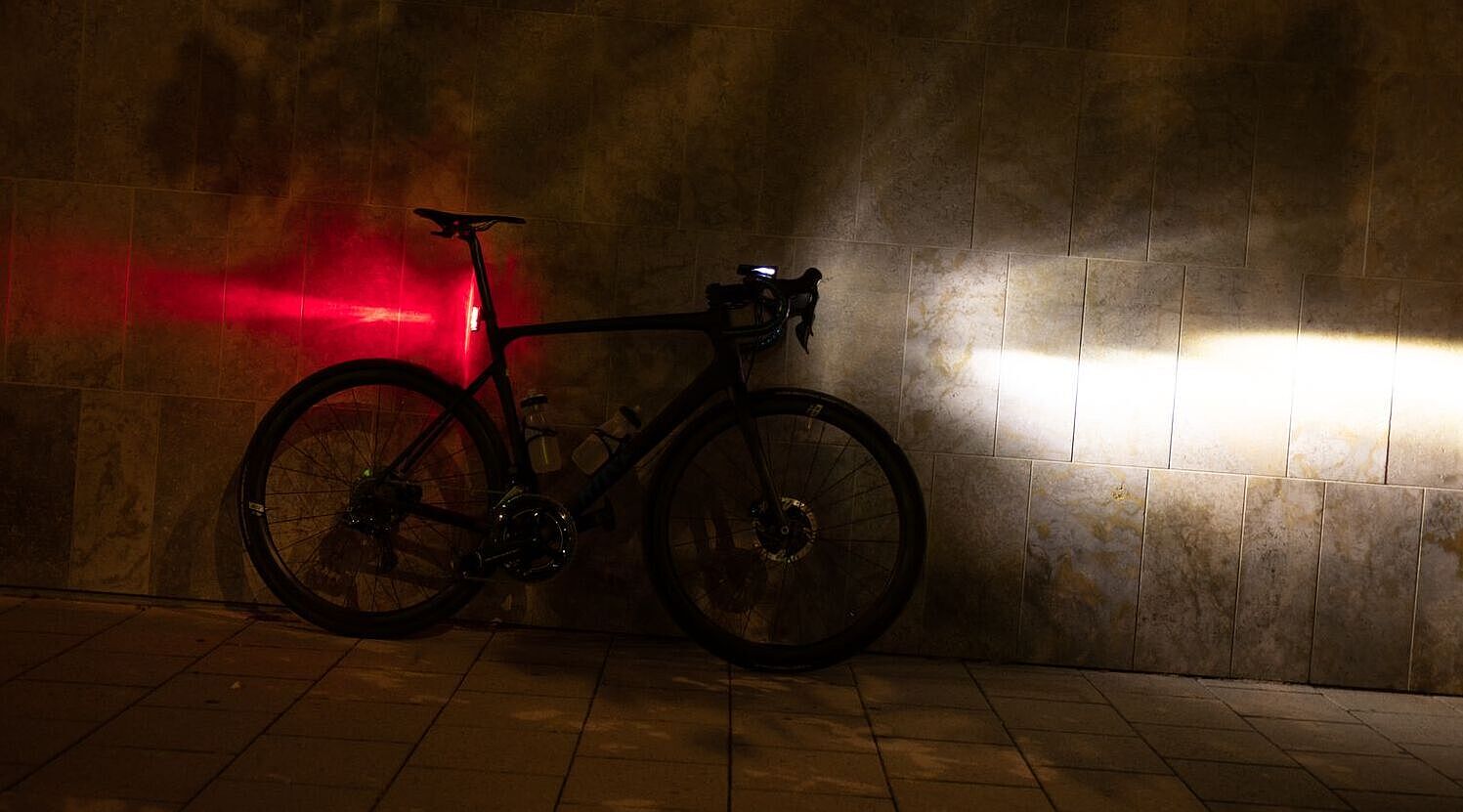 Ein Fahrrad steht in der Dunkelheit an einer Wand, mit am Lenker montiertem, eingeschalteten Scheinwerfer und an der Sattelstütze montierten, leuchtenden Rücklicht.