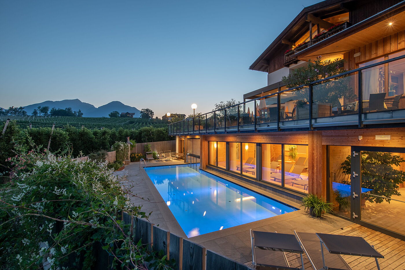 Sommerabende am Pool Außenbereich eines modernen Hotels zur blauen Stunde; im Vordergrund ein beleuchteter Swimmingpool und Liegestühle auf einer Holzterrasse, im Hintergrund Weinberge und eine Bergkette unter dem Abendhimmel.