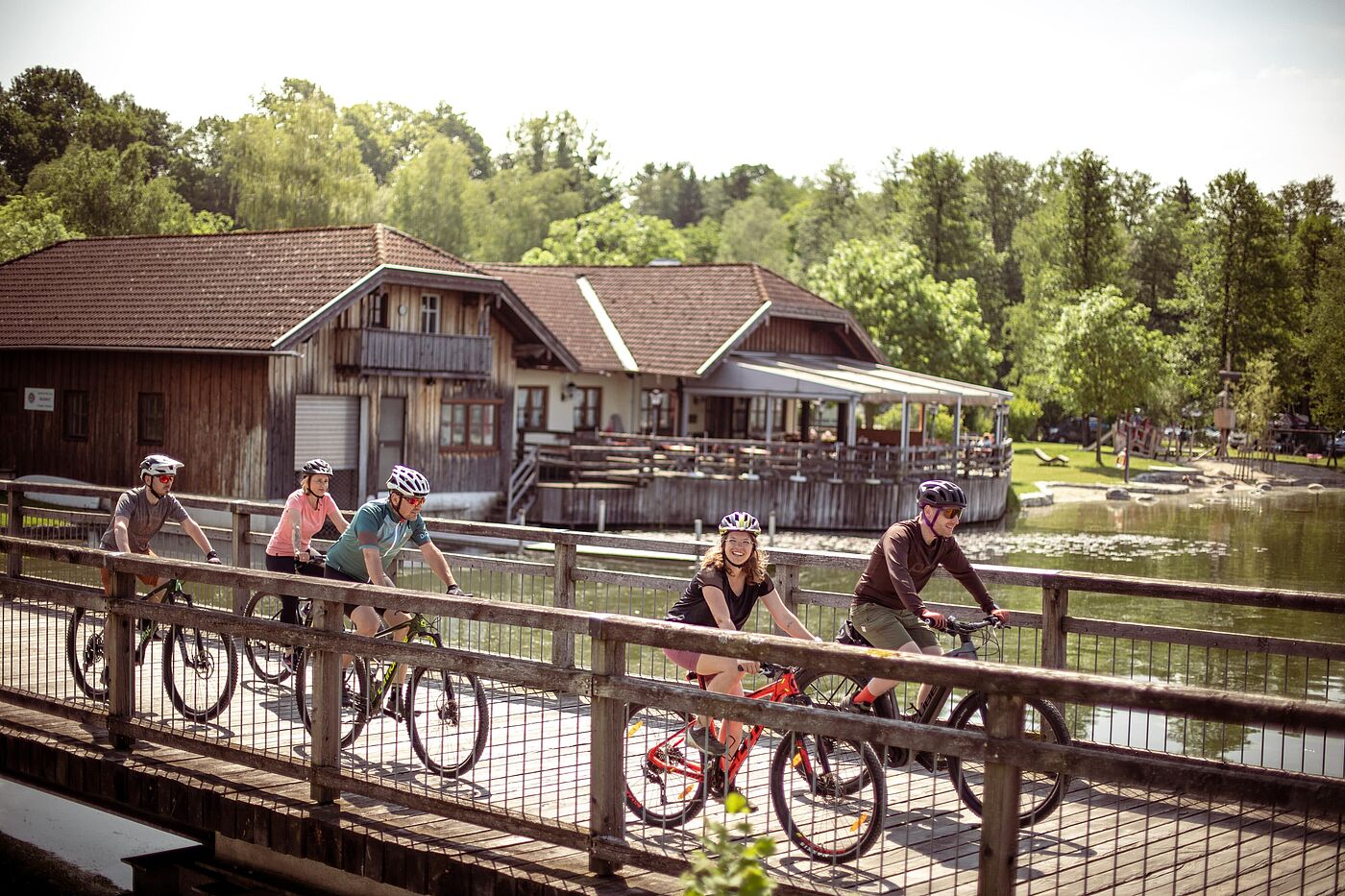 Fünf Personen fahren mit Fahrrädern über eine Holzbrücke an einem See entlang.