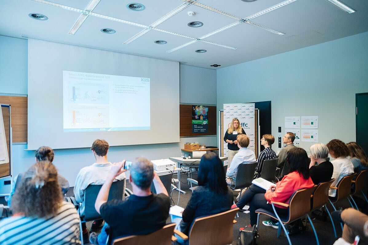 In einem Raum präsentiert eine Frau mithilfe einer Wandprojektion. Im Vordergrund ist Publikum sichtbar. 