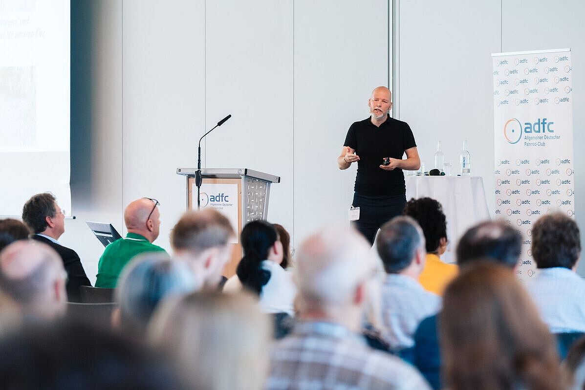 Dr. Stefan Carsten hält auf einer Bühne einen Vortrag. Im Vordergrund ist Publikum zu sehen. Im Hintergrund ein Rednerpult, weiße Wand und ein weißer Aufsteller mit ADFC-Logos. 