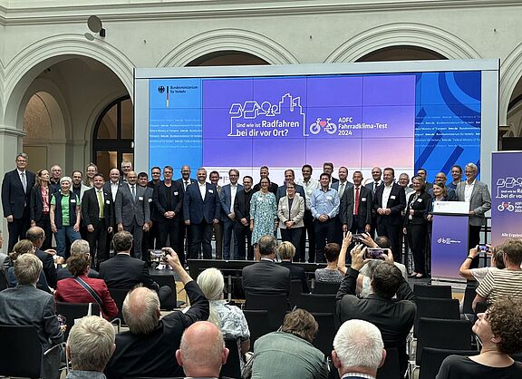 Auf einer großen Bühne im Bundesverkehrsministerium sind zahlreiche Städtevertreterinnen und -vertreter versammelt, eingerahmt von Bundesverkehrsminister Schnieder und ADFC-Bundesvorsitzenden Masurat. 