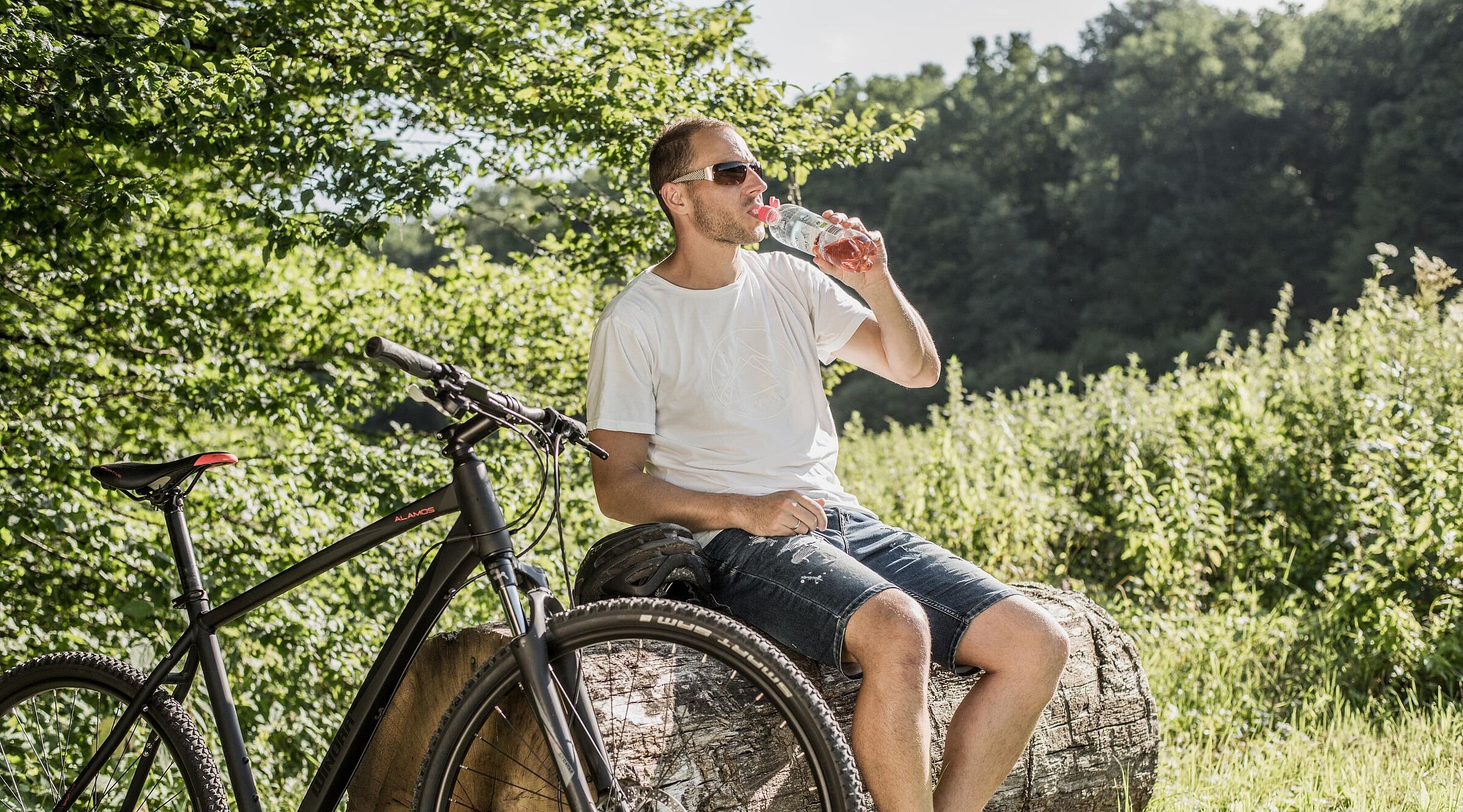 Radfahrer mit Sonnenbrille trinkt Wasser aus einer Flasche während einer Pause auf einem Baumstamm. Sein schwarzes Mountainbike steht daneben.