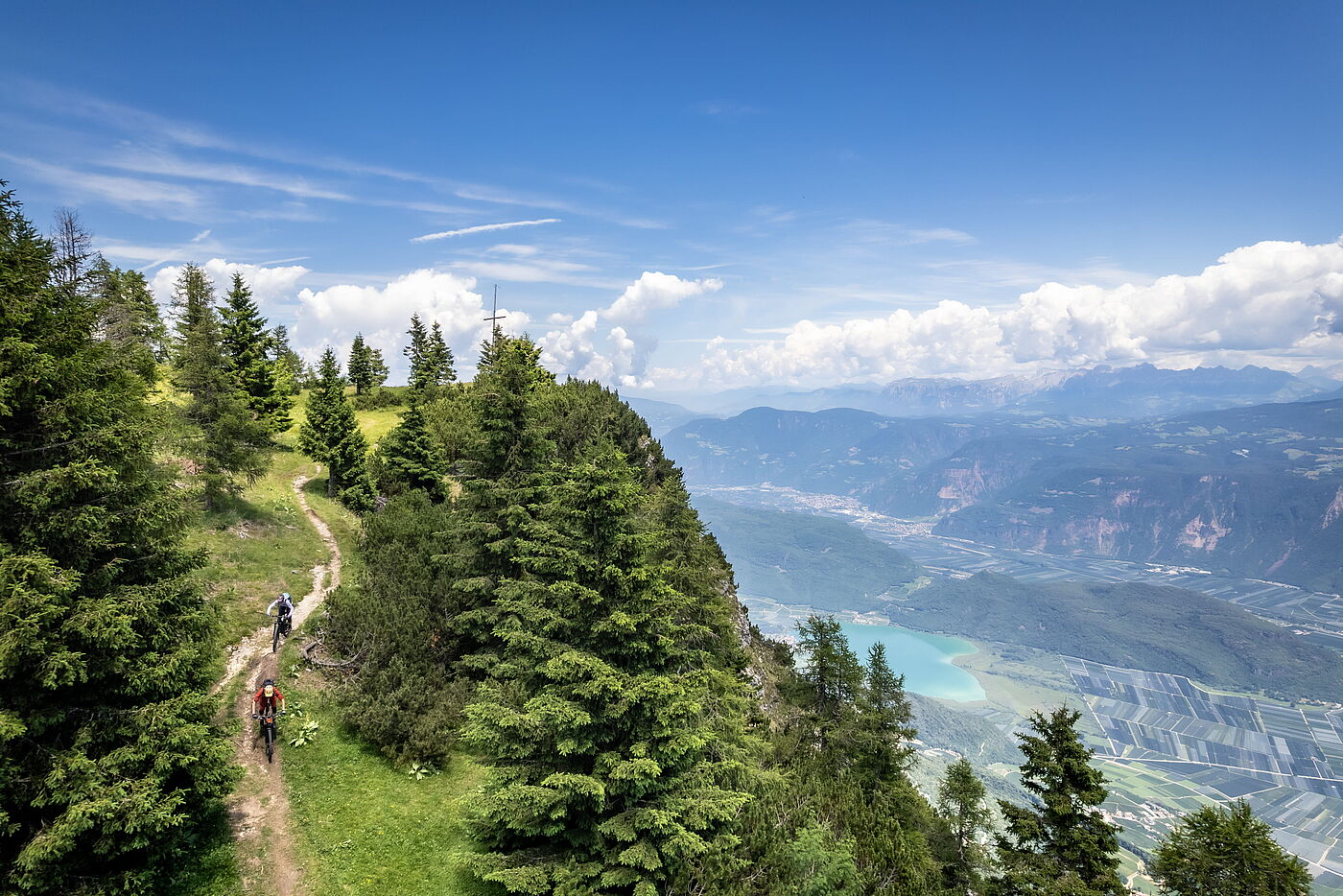 Zwischen Gipfelglück und Talblick Zwei Mountainbiker fahren auf einem schmalen Waldpfad entlang eines Berghangs; rechts fällt der Blick steil hinab auf einen türkisfarbenen See im Tal und weite Obstplantagen.