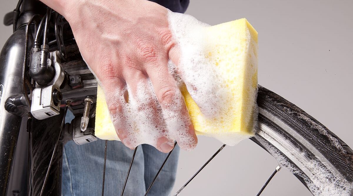 Großaufnahme einer Hand mit Schwamm und Schaum, die ein Fahrrad-Vorderrad säubert. 