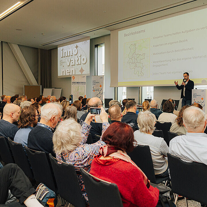 Viele Menschen sitzen in einem Saal und hören zwei Personen zu, die auf der Bühne des ADFC Kongress 2026 stehen. Eine große Leinwand zeigt Folien zum Vortrag.