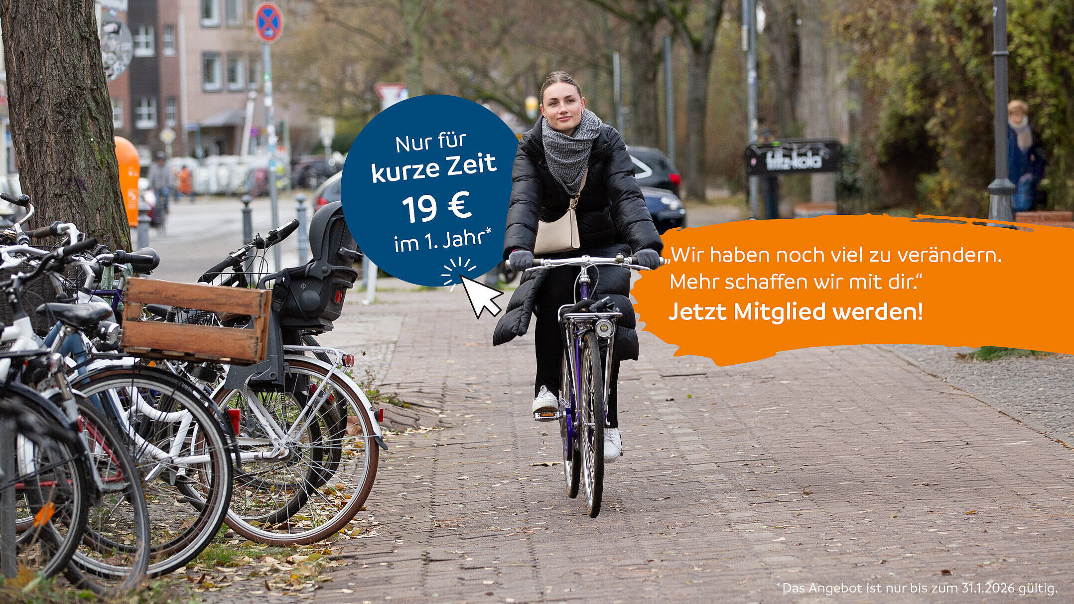 Eine Frau fährt auf einem Fahrrad einen gepflasterten Weg entlang, vorbei an abgestellten Rädern und herbstlichen Bäumen.