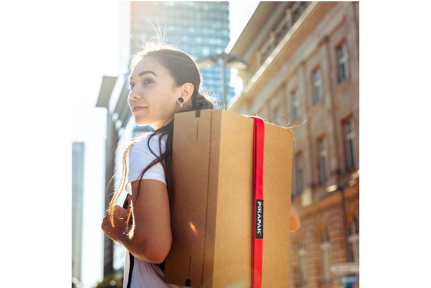 Pikapak Eine Person geht draußen in der Sonne und trägt ein rechteckiges Paket mit rotem Gurt auf dem Rücken