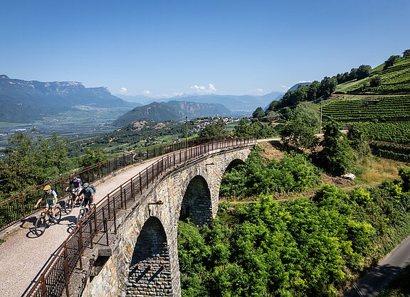 Auf alten Pfaden zu neuen Aussichten Drei Mountainbiker fahren bei sonnigem Wetter über eine historische Steinbogenbrücke; rechts erstrecken sich grüne Weinterrassen, im Hintergrund liegt ein weites Tal vor einer Bergkette.