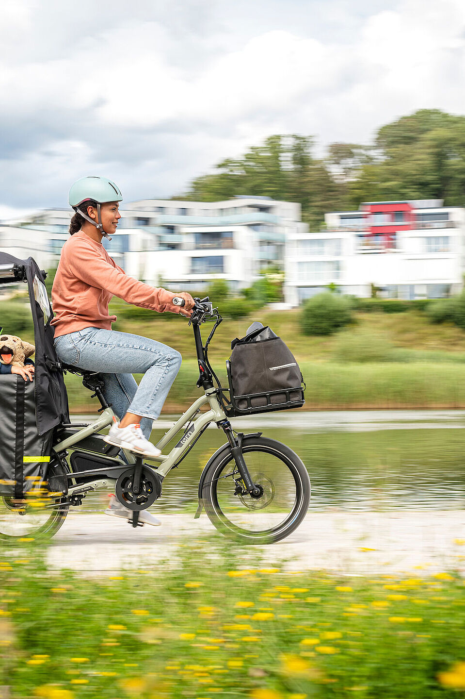 Tern Zubehör Eine junge Frau fährt auf einem Longtail mit Kind als Passagier, das in einem wetterfesten Aufbau sitzt.