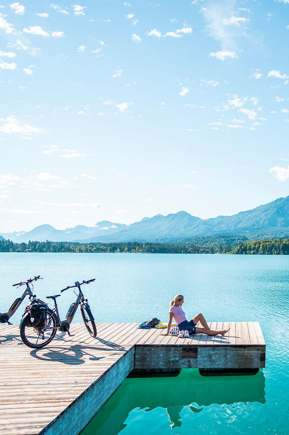 Badepause am Bergsee nach der Radtour Person springt von Holzsteg ins Wasser, Elektrofahrräder stehen daneben, Bergpanorama im Hintergrund.