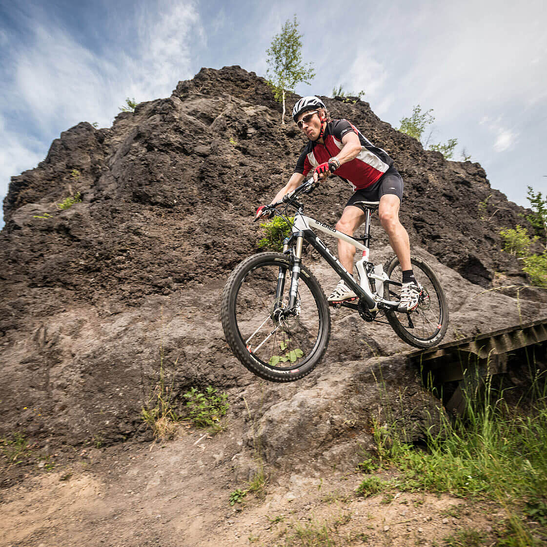 Mountainbike Action in der Eifel Ein Mountainbiker fährt über einen steinigen Weg und eine kleine Holzbrücke. Hinter ihm ragt ein dunkler Felsen auf.