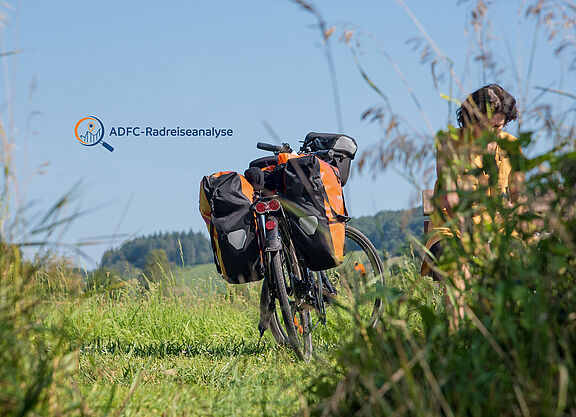 Reiserad mit Packtaschen steht in einer gemähten Wiese, Frau hinter hochgewachsenem Gras