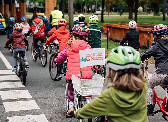 Mehrere Kinder und Erwachsene fahren bei einer Kidical Mass gemeinsam mit Fahrrädern und Helmen auf einer Straße. Ein Kind trägt eine Weste und hat ein Schild mit „Kidical Mass“ am Fahrradkorb.