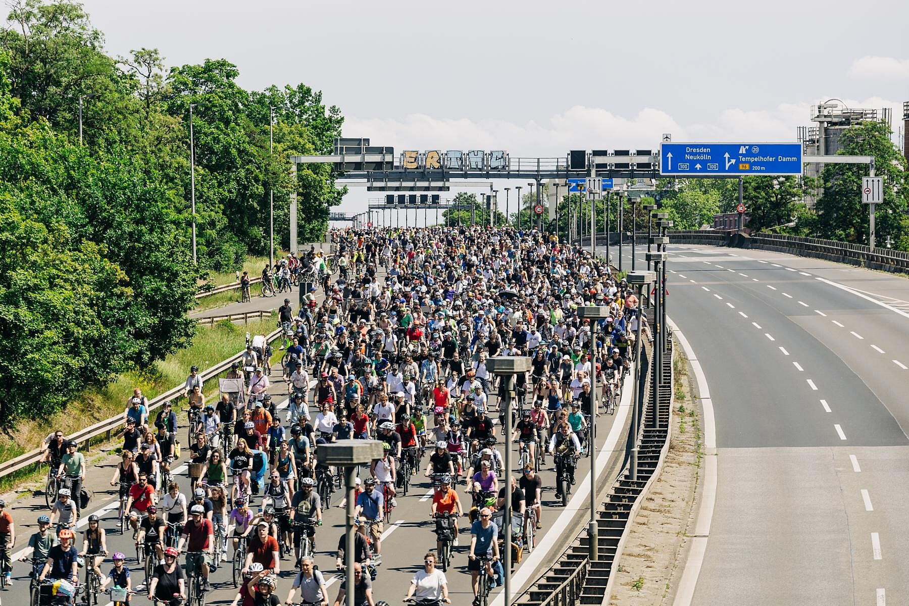 Eine große Menschenmenge fährt auf Fahrrädern über eine mehrspurige Autobahn, die bis zum Horizont mit Radfahrenden gefüllt ist. Über der Straße befinden sich Verkehrsschilder, die unter anderem Richtung „Dresden“ und „Neukölln“ zeigen. Links säumen Bäume die Straße, rechts ist eine leere Gegenfahrbahn zu sehen.