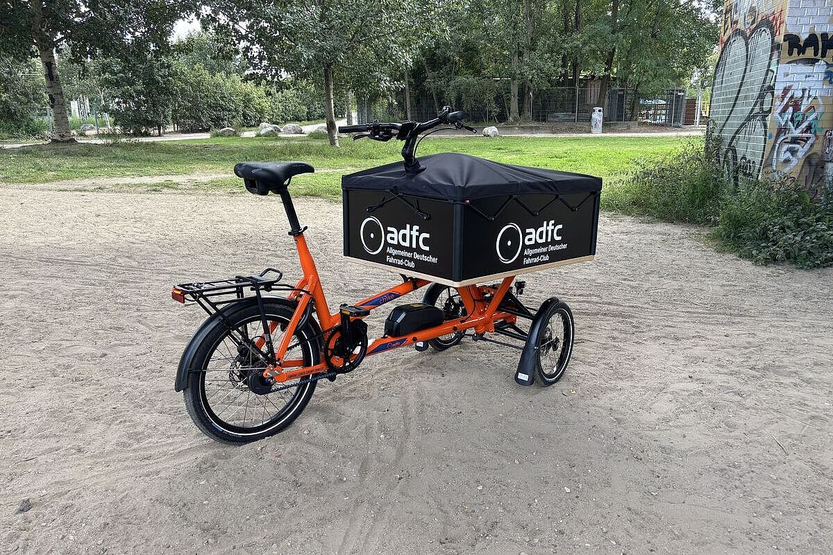 Chike ADFC Orangefarbenes dreirädriges Lastenrad mit schwarzer ADFC-Transportbox steht auf einem sandigen Weg im Grünen.