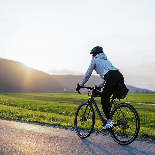 Eine Person fährt mit dem Fahrrad auf einer ruhigen Straße durch eine weite Wiesenlandschaft, die Sonne steht tief am Himmel.