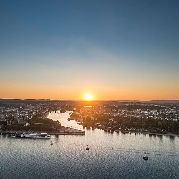 Luftaufnahme von Koblenz bei Sonnenuntergang. Im Vordergrund fließt ein breiter Fluss, darüber schweben mehrere Seilbahngondeln.