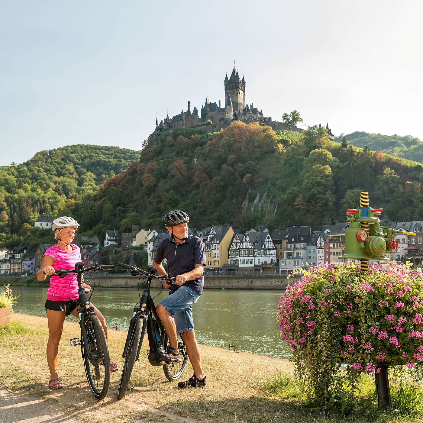 Radfahren auf dem Mosel-Radweg bei Cochem Eine Frau und ein Mann stehen mit Fahrrädern an der Mosel. Hinter ihnen liegen bunte Häuser und die Reichsburg Cochem auf einem bewaldeten Hügel.