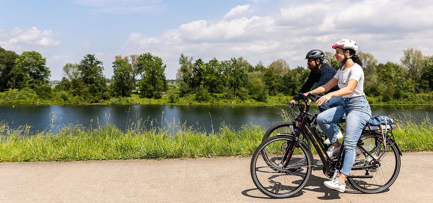 Radfahrende auf der Ikonen-Route Zwei Radfahrer mit Helmen fahren auf einem asphaltierten Radweg entlang eines Sees oder Flusses. Im Hintergrund sind grüne Bäume und eine natürliche Uferlandschaft zu sehen.