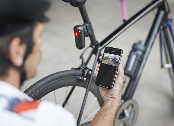 Mann kniet mit Smartphone in der Hand vor einen Rennrad mit rot leuchtendem Rücklicht an Sattelstütze. Das Rücklicht Garmin Varia RCT715 mit integrierter Kamera zeichnet permanent auf und speichert Daten aber nur bei einem erkannten Unfall.  