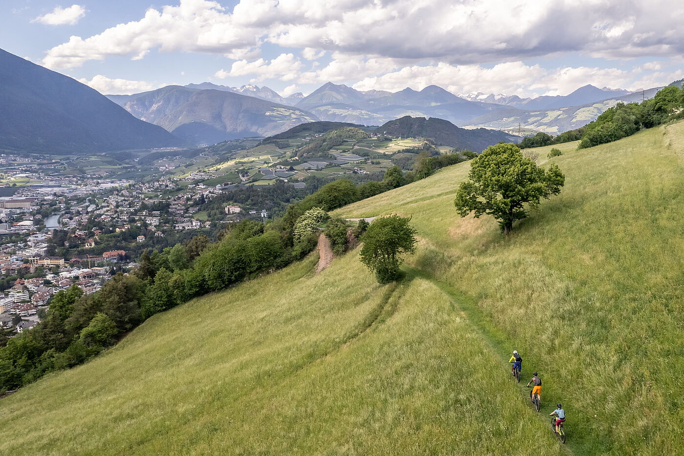 Freiheit über den Dächern Drei Mountainbiker fahren hintereinander auf einem schmalen Singletrail über eine grüne Almwiese; links unten liegt eine Stadt im Tal, dahinter erstreckt sich eine weite Bergkette unter bewölktem Himmel.