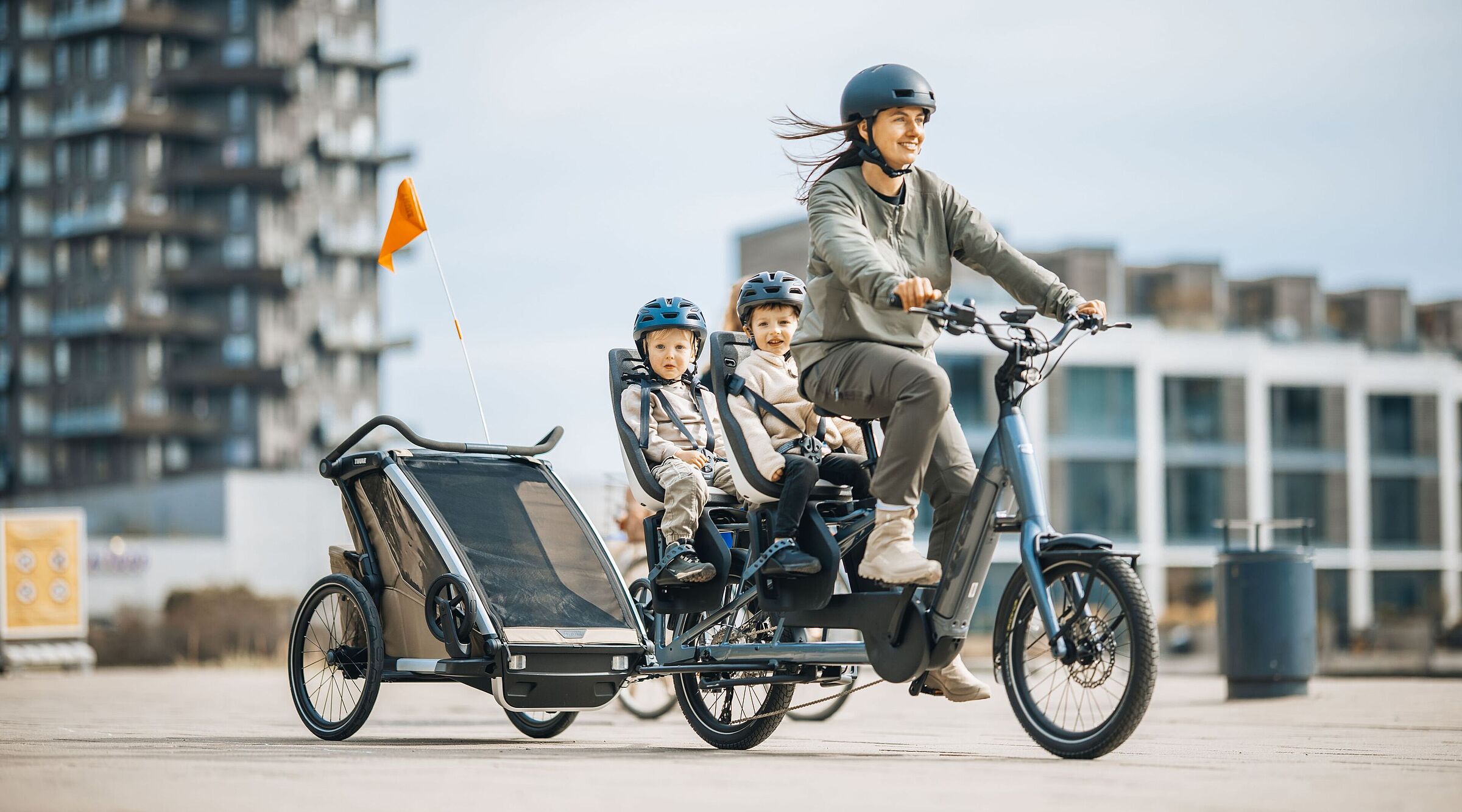 Frau fährt ein Lastenrad Longtail, auf dem langen Gepäckträger hinten sitzen zwei Kinder in je einem Kindersitz. Das Fahrrad zieht zudem einen Kinderfahrradanhänger.