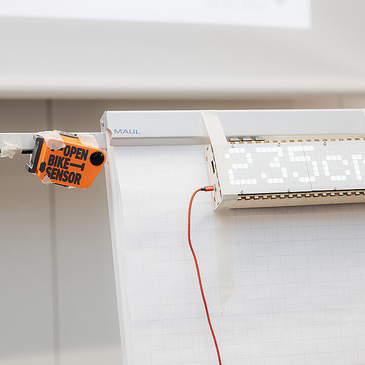 Ein orangefarbener Open Bike Sensor und ein digitales Display mit der Anzeige 235 cm sind an einem Flipchart befestigt.